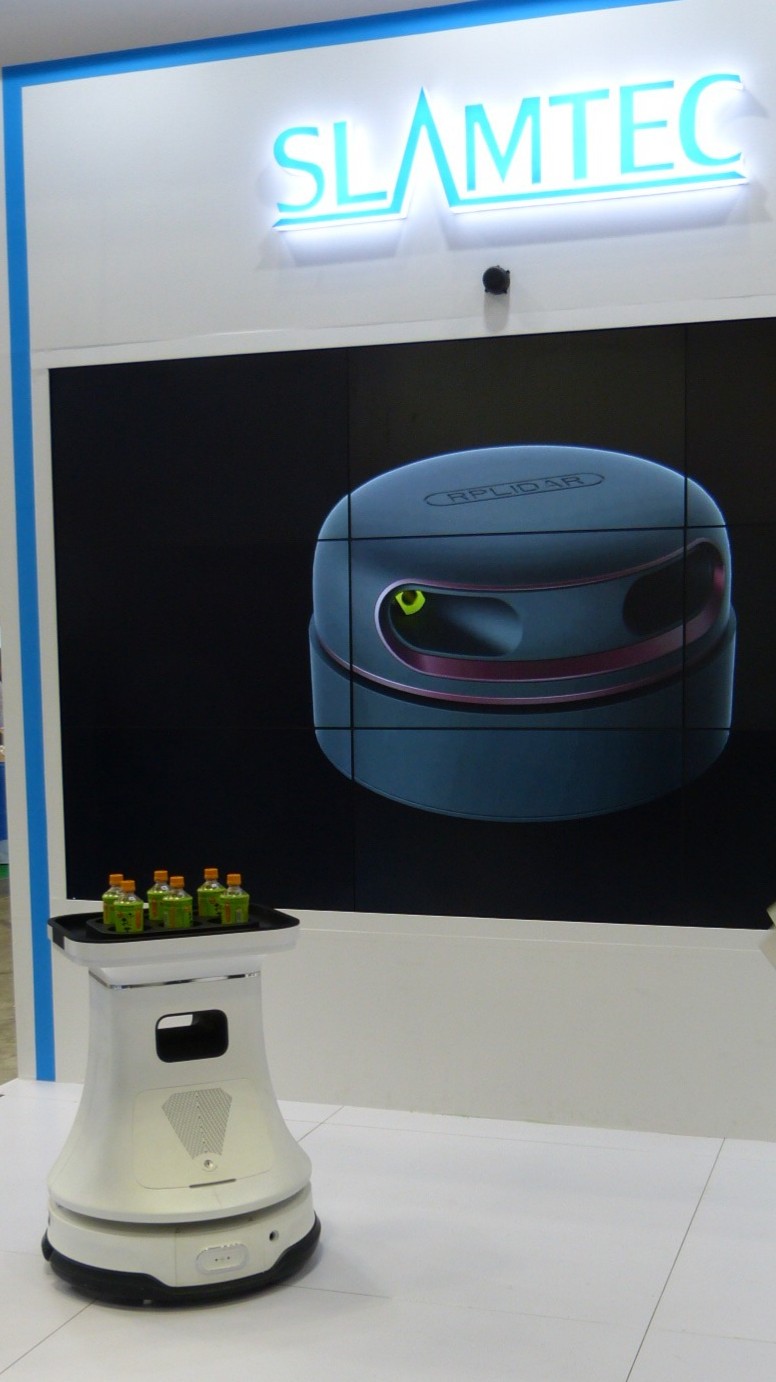 a waist high robot similar to a roomba supports a tray of bottled drinks near a Slamtec logo